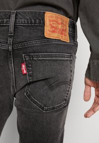 Jeanși negri din denim cu o textură estompată, având o etichetă din piele maro deschis pe care este inscripționat "Levi Strauss & Co." și un tag roșu Levi's pe buzunar.