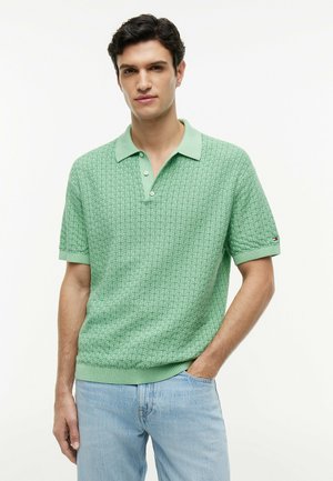 Mand iført lysegrøn tekstureret kortærmet poloshirt og lyseblå jeans, stående med den ene hånd i lommen mod en hvid baggrund.