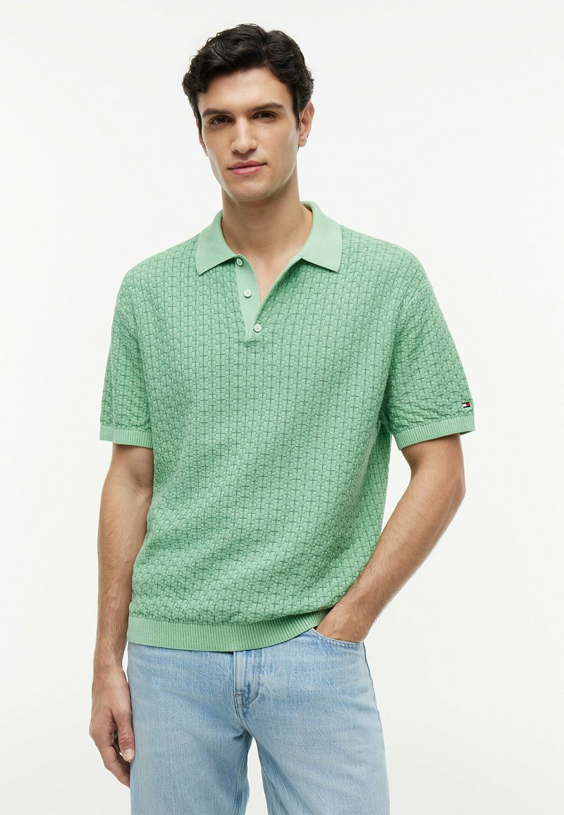 Homme portant un polo vert clair texturé à manches courtes et un jean bleu clair, debout avec une main dans la poche sur fond blanc.