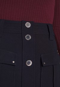 Falda azul marino con pliegues, con cierres de botones plateados y bolsillos frontales. Hecha de tela texturizada y estructurada. Parte superior burdeos parcialmente visible.