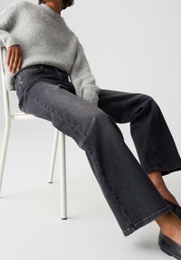 Jeans noirs à jambes larges avec une texture douce et une décoloration subtile. Associés à un pull côtelé gris clair. Modèle assis sur une chaise blanche.