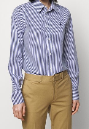 Homme portant une chemise à rayures verticales bleues et blanches à manches longues, rentrée dans un pantalon beige, sur un fond uni.