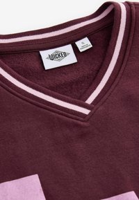 Burgundy V-ringad sweatshirt med rosa randig kant, mjuk textur, stor ljusrosa grafisk detalj och "Wicked"-etikett inuti kragen.