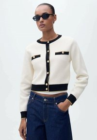 Cardigan crème avec des accents noirs, des boutons dorés et deux poches avant. Présente des poignets et un ourlet côtelés. Associé à un denim bleu foncé.