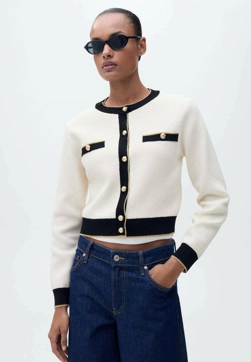 Cardigan crème avec des accents noirs, des boutons dorés et deux poches avant. Présente des poignets et un ourlet côtelés. Associé à un denim bleu foncé.