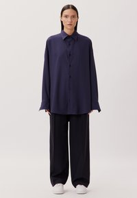 Marineblaue, oversized Knopfleiste-Hemd mit einem klaren Kragen, langen Ärmeln und einer glatten Textur, kombiniert mit weiten, dunklen Hosen und weißen Sneakers.