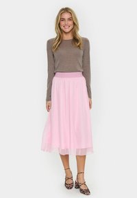 Lunga gonna rosa realizzata in tessuto leggero, con vita arricciata e design fluido, abbinata a una maglietta marrone a maniche lunghe aderente e scarpe a fantasia.