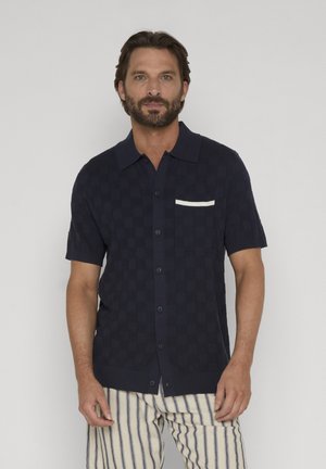 Mann med skjegg iført en marineblå kortermet skjorte med knapper, hvit kant ved lomma, og beige bukser med blå vertikale striper.