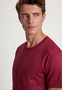 Burgunderrotes Kurzarm-T-Shirt aus einem weichen Stoff, mit einem runden Ausschnitt und einer aufgesetzten Tasche auf der linken Brust. Glatte Textur.