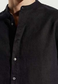 Primer plano de una persona que lleva una camisa negra de cuello mandarín con botones visibles y parte del cuello y el pecho.