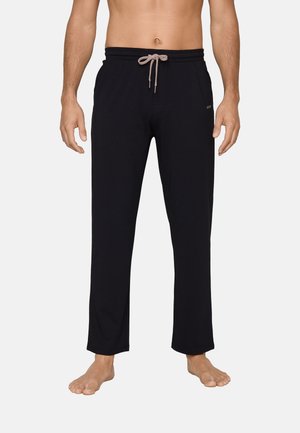 Man draagt zwarte broek met rechte pijpen en een lichtgrijze trekkoorden, staat op blote voeten tegen een effen achtergrond.