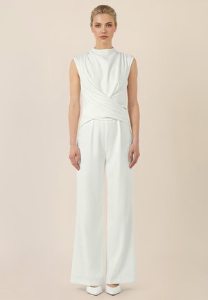 Kobieta o blond włosach w białym topie bez rękawów z zakładką i szerokich białych spodniach stoi na tle jednolitego beżowego tła.