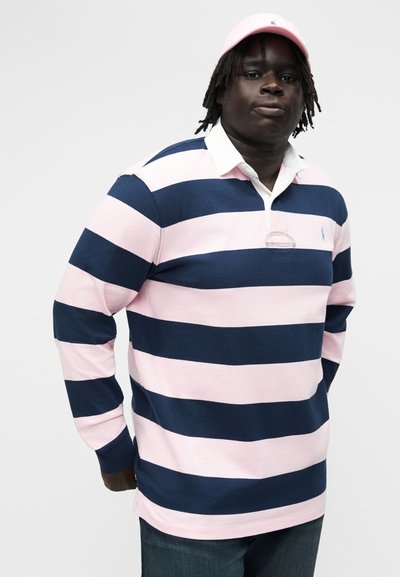 Mann iført navyblå og lyserosa rugbyskjorte med horisontale striper og lyserosa baseballcaps, stående mot en ensfarget lys bakgrunn.