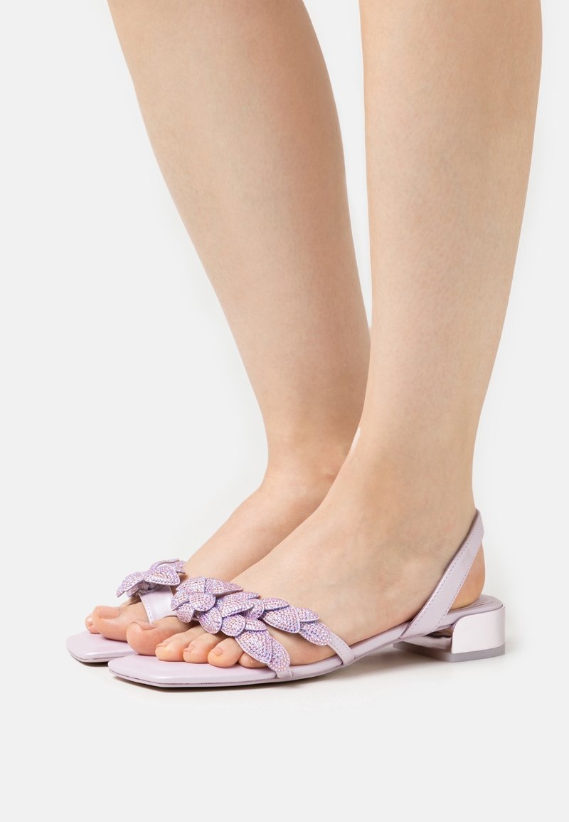 Pieds portant des sandales ouvertes violet clair avec de petits talons blocs et des ornements décoratifs en forme de feuille sur les brides.