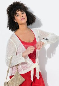 Cardigan bianco in maglia con fronte legato, indossato su una maglietta rossa con scollatura a scallop e pois, abbinato a una borsa d'oro testurizzata.