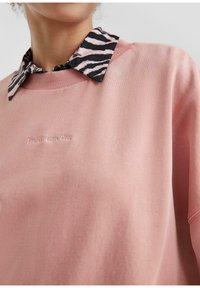Persona vestida con un sudadera rosa con "The 05 Love Club" bordado y una camisa negra con estampado de cebra rosa debajo.