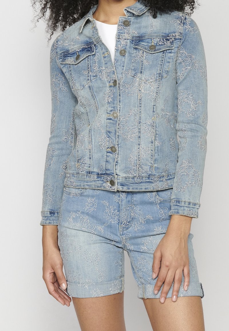 Femme portant une veste en jean bleu clair et un short avec une broderie florale blanche, debout avec la main relâchée le long du corps.