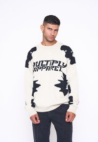 Pulover crem cu modele de frunze negre și textul "MULTIPLY APPAREL". Are mâneci lungi și manșete ondulate. Purtați-l cu pantaloni negri.