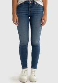 Kitsad teksased tumedas sinistes denimis, kõrge talje ja kerged kulumisjäljed, koos valgete spordijalatsitega. Omab heledat õmblust ja viit taskut.