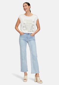 Chemisier sans manches en coton blanc avec un texte estival, associé à un jean évasé bleu clair. Le modèle porte des sandales à plateforme beiges. Design simple.