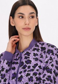 Giacca bomber viola con stampa animale nera, dotata di chiusura con zip e polsini e colletto a coste. Il tessuto testurizzato aggiunge interesse visivo.