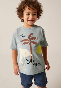 Lyseblå t-shirt med korte ærmer, der viser en delfin, et palmetræ, solen og teksten "Sun Vibes" i kontrasterende farver; blødt stof, rund hals.