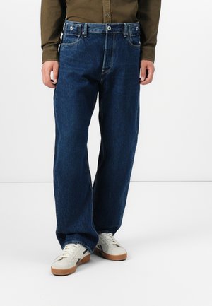Dark blue denim jeans with a loose fit, featuring a button and zipper closure, five-pocket design, and subtle fading. Paired with white sneakers.