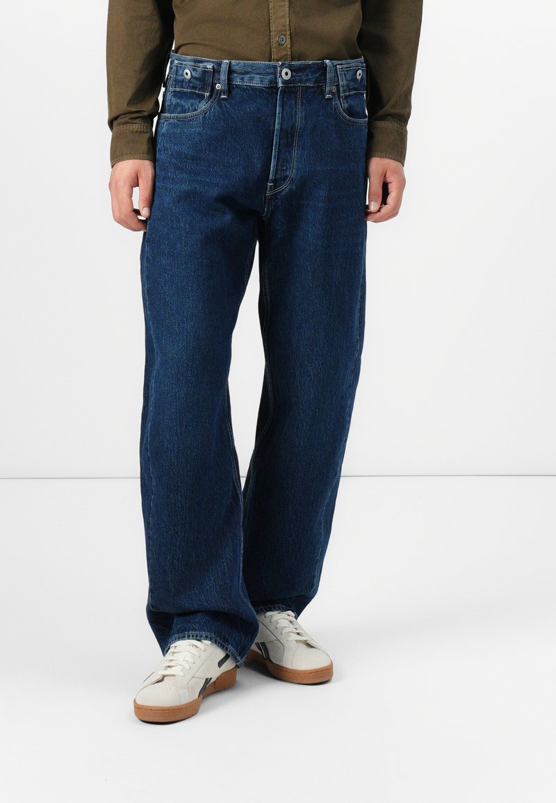 Jean en denim bleu foncé à coupe ample, avec une fermeture à bouton et à zip, un design à cinq poches et un léger délavage. Associé à des baskets blanches.