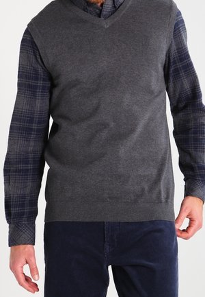 Mies, joka on pukeutunut tummanharmaaseen hihattomaan V-kaulaiseen neuleeseen, jonka alla on laivastonsininen ruudullinen paita ja tummansiniset housut, kädet rentona sivuilla.