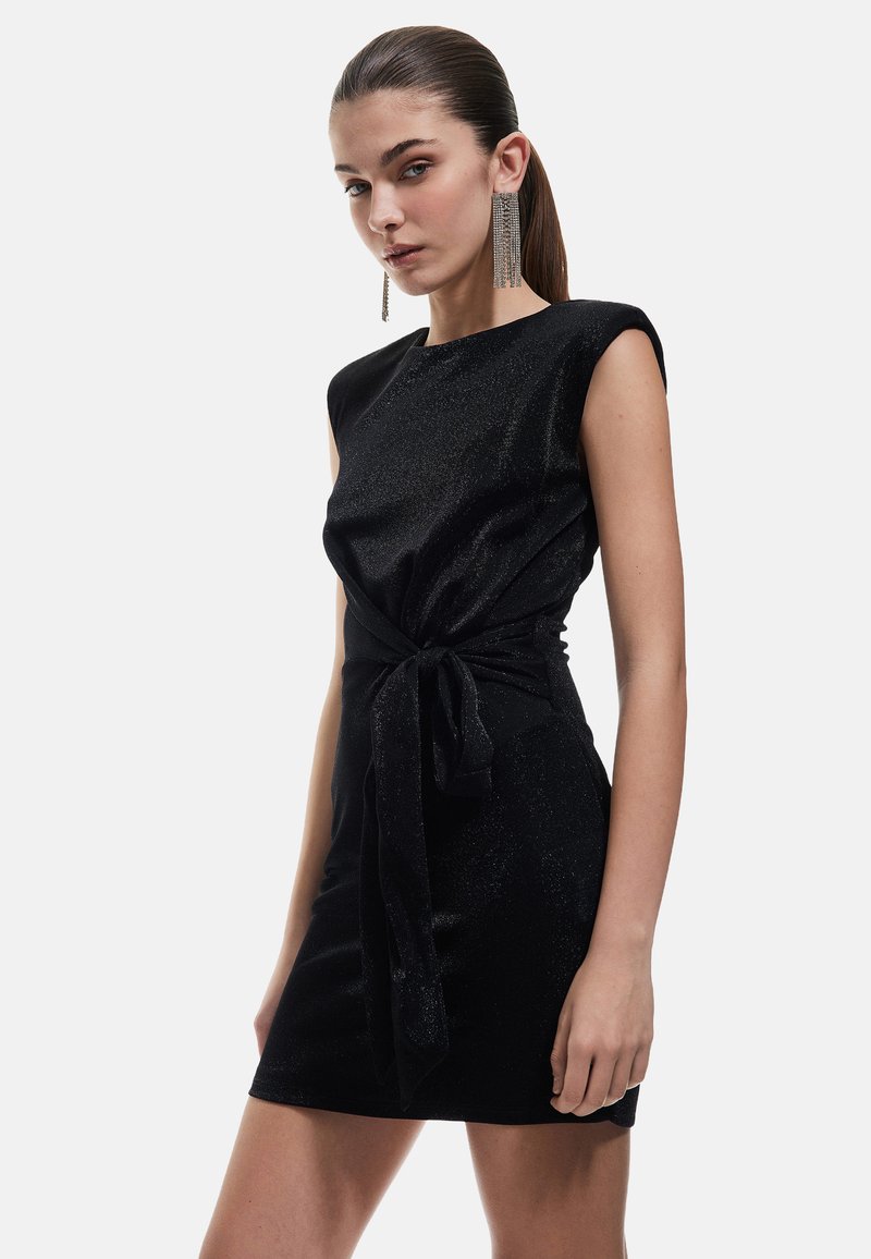 Robe noire courte à la coupe ajustée, avec des manches courtes, un détail de ceinture à la taille et une texture légèrement brillante, complétée par des boucles d'oreilles pendantes.