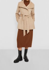 Beige jas met ceintuur en brede kraag, gecombineerd met een bruine geribde midi-rok en glanzende zwarte enkellaarzen.