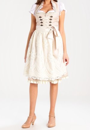 Femme portant une robe traditionnelle beige avec un tablier en dentelle, des manches blanches bouffantes et des talons beiges, debout sur un fond uni.