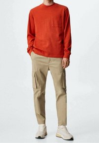 Langsleeve, ronde hals, oranje shirt met een voorzak; gecombineerd met beige cargobroek met zijzakken; witte sneakers.