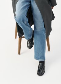 Black leather loafers with tassel detail, worn with light blue jeans, seated on a wooden chair, against a plain white background.