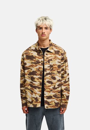 Junger Mann mit kurz gebleichtem Haar, der eine braun-beige Tarnjacke über einem schwarzen Hemd und grauen Hosen trägt, steht vor einem einfarbigen Hintergrund.
