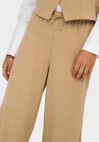 Pantalons larges beige en tissu léger et texturé. Dotés d'une taille élastique avec cordon de serrage et de poches avant.