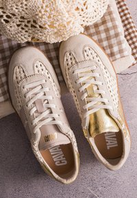 Paire de baskets beige et or avec des détails tissés et des lacets, placées sur un tissu plié à carreaux et uni sur une surface texturée.