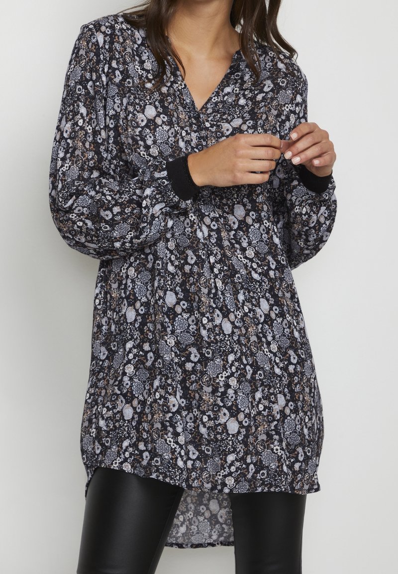 Femme portant une tunique noire à manches longues avec motif floral et poignets noirs, portée sur des leggings en cuir noirs, sur un fond uni.