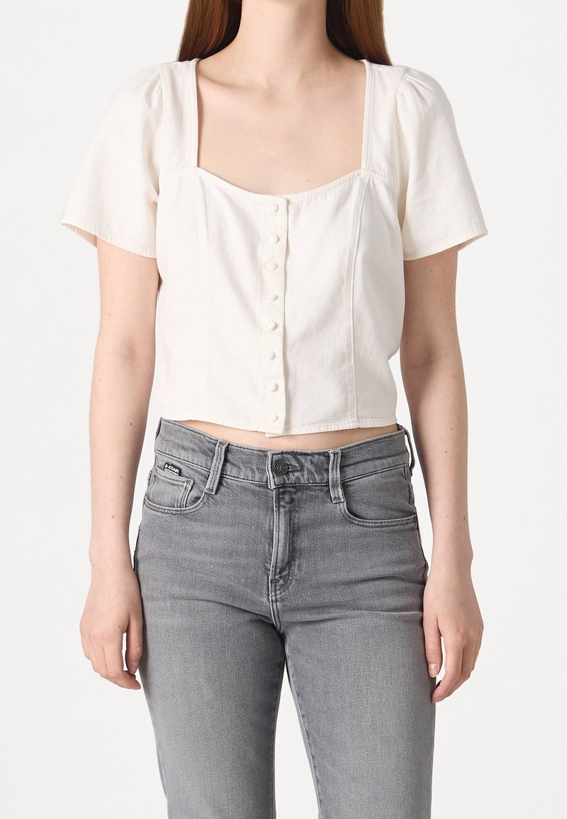 Top corto bianco a maniche corte con scollo quadrato e chiusura a bottoni sul davanti; abbinato a jeans in denim grigio a vita alta.