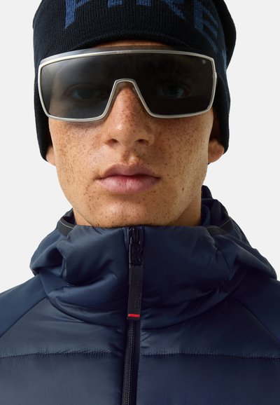 Marineblaue Steppjacke mit hohem Kragen und Reißverschluss, schwarze Strickmütze und große rechteckige Sonnenbrille mit silbernen Rahmen. Sichtbare strukturierte Haut.