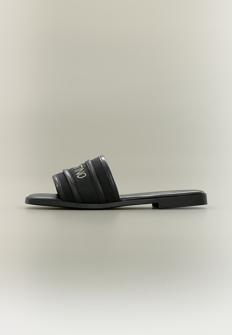 Sandale plate noire à bride large avec trois brides rembourrées sur le dessus du pied et un petit talon, présentée sur un fond neutre.