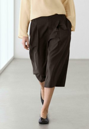 Persona che cammina al chiuso indossando pantaloni corti larghi marrone scuro, maglia giallo pallido a manica lunga e scarpe basse nere.