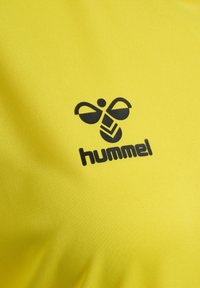 Żółta sportowa koszulka wykonana z teksturowanej tkaniny, z czarnym logo o wzorze przypominającym owada oraz napisem „hummel” poniżej.