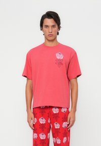 Camiseta de algodón roja con cuello redondo y mangas cortas, que presenta un gráfico de una rosa rosa en el lado izquierdo. Modelo vistiendo pantalones con estampado floral.