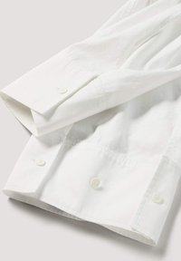 Witte katoenen blouse met lange mouwen, voorzien van knopen op de manchetten en een gladde structuur. Opgevouwen presentatie toont strakke lijnen en een verzorgd ontwerp.