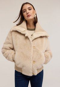 Veste en fausse fourrure beige avec col côtelé, fermeture à boutons, poches latérales et finition texturée. Portée sur un pull vert et un jean.