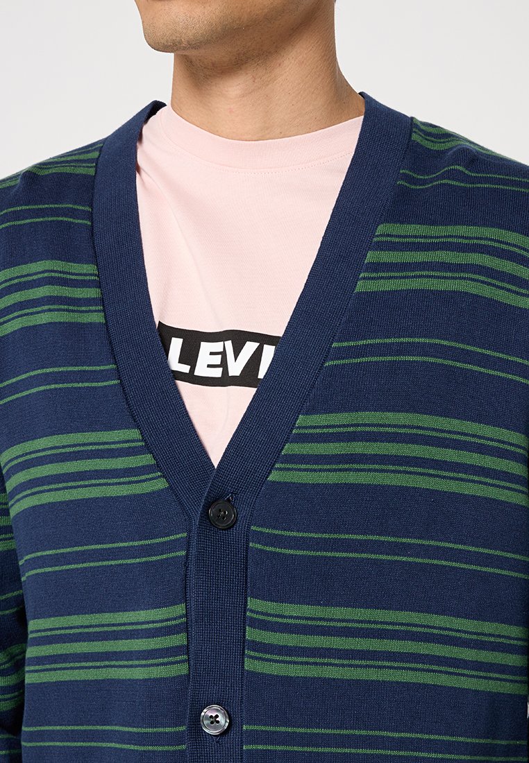 Man in een marineblauwe cardigan met groene strepen over een lichtroze Levi's T-shirt, met knopen aan de voorkant.