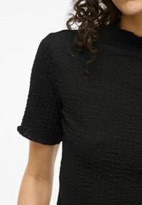 Maglia nera a texture con maniche corte, caratterizzata da un motivo a coste e un collo alto. Il tessuto appare morbido e leggermente stropicciato.