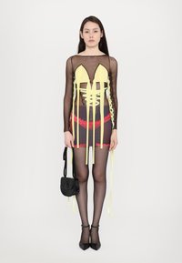 Gelbes und schwarzes durchscheinendes Kleid mit Schnürdetails, roten Spitzenakzenten, kombiniert mit schwarzen High Heels und einer kleinen schwarzen Tasche.