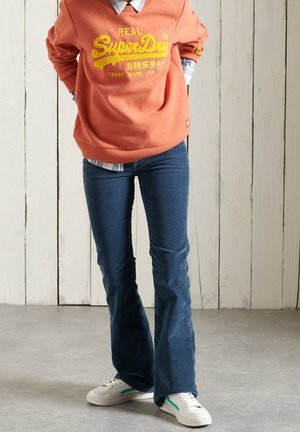 Korálová mikina se žlutým logem, modré rozšířené džíny a bílé tenisky se zelenými detaily. Pozadí tvoří světlé dřevěné stěny.